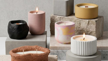 Modern concrete candle jars in various shapes and colors, displayed on stone blocks with soft burning flames.